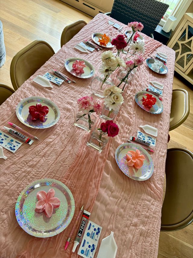 Indoor home dining table set for a pink DIY craft party: long textured pink tablecloth, holographic plates with flower-shaped decorations, glue tubes, small trays and tools at each place, and mixed-flower glass-vase centerpieces.
