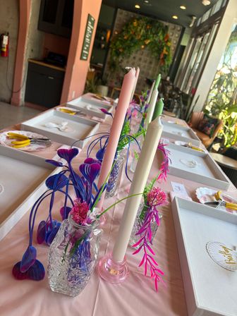 Craft workshop table in a bright indoor studio set for a candle-making party — pink and cream taper candles in glass holders, vibrant purple and pink faux florals, trays with tools on a blush tablecloth.