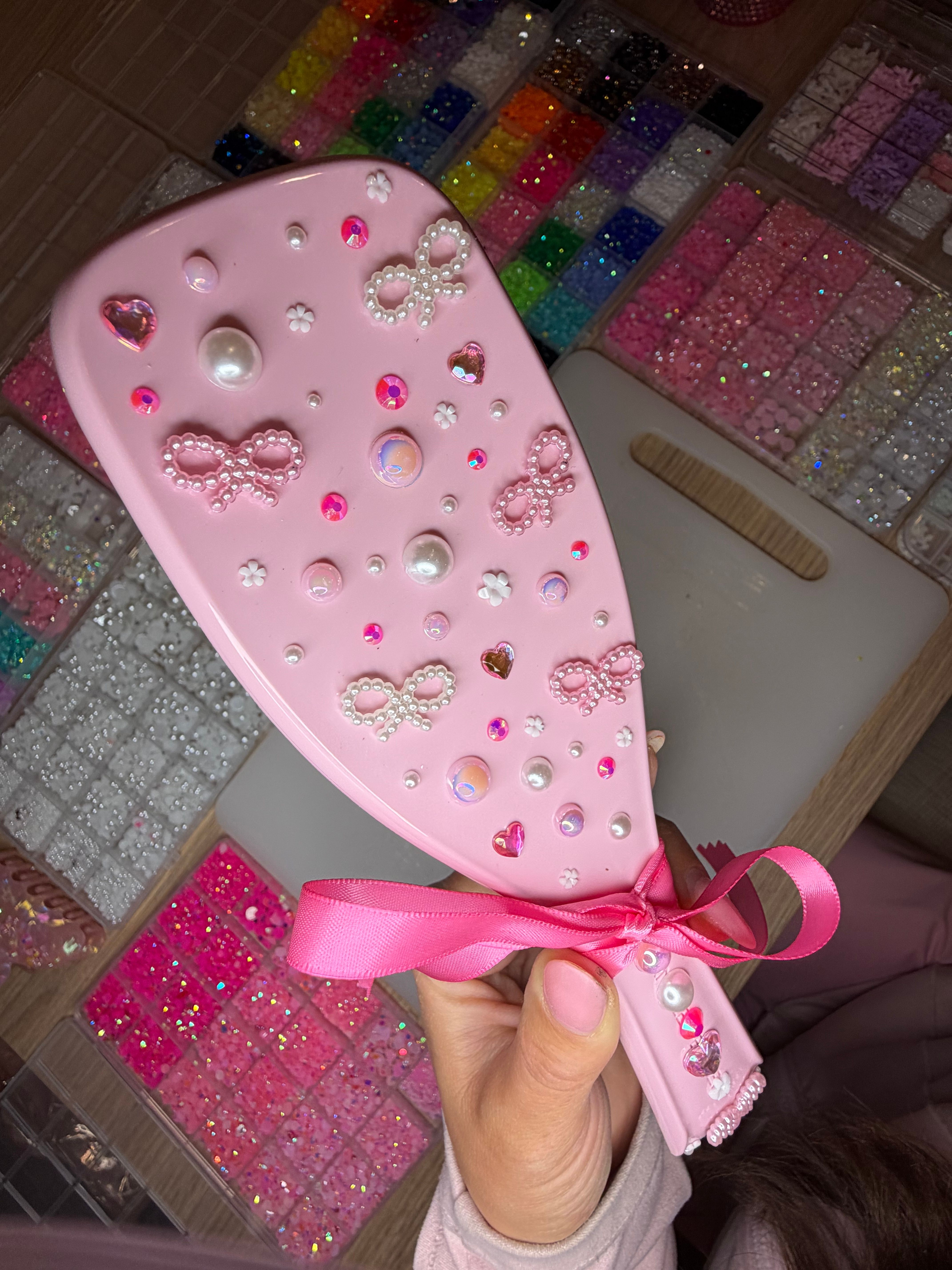 Pink handheld hairbrush decorated with pearl bow embellishments, heart rhinestones, pearls and iridescent gems, held over trays of colorful glitter beads for DIY crafts