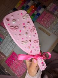 Pink handheld hairbrush decorated with pearl bow embellishments, heart rhinestones, pearls and iridescent gems, held over trays of colorful glitter beads for DIY crafts