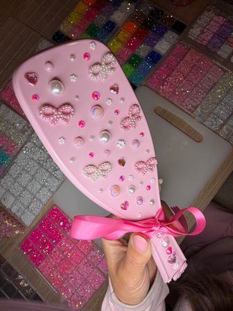 Pink handheld hairbrush decorated with pearl bow embellishments, heart rhinestones, pearls and iridescent gems, held over trays of colorful glitter beads for DIY crafts