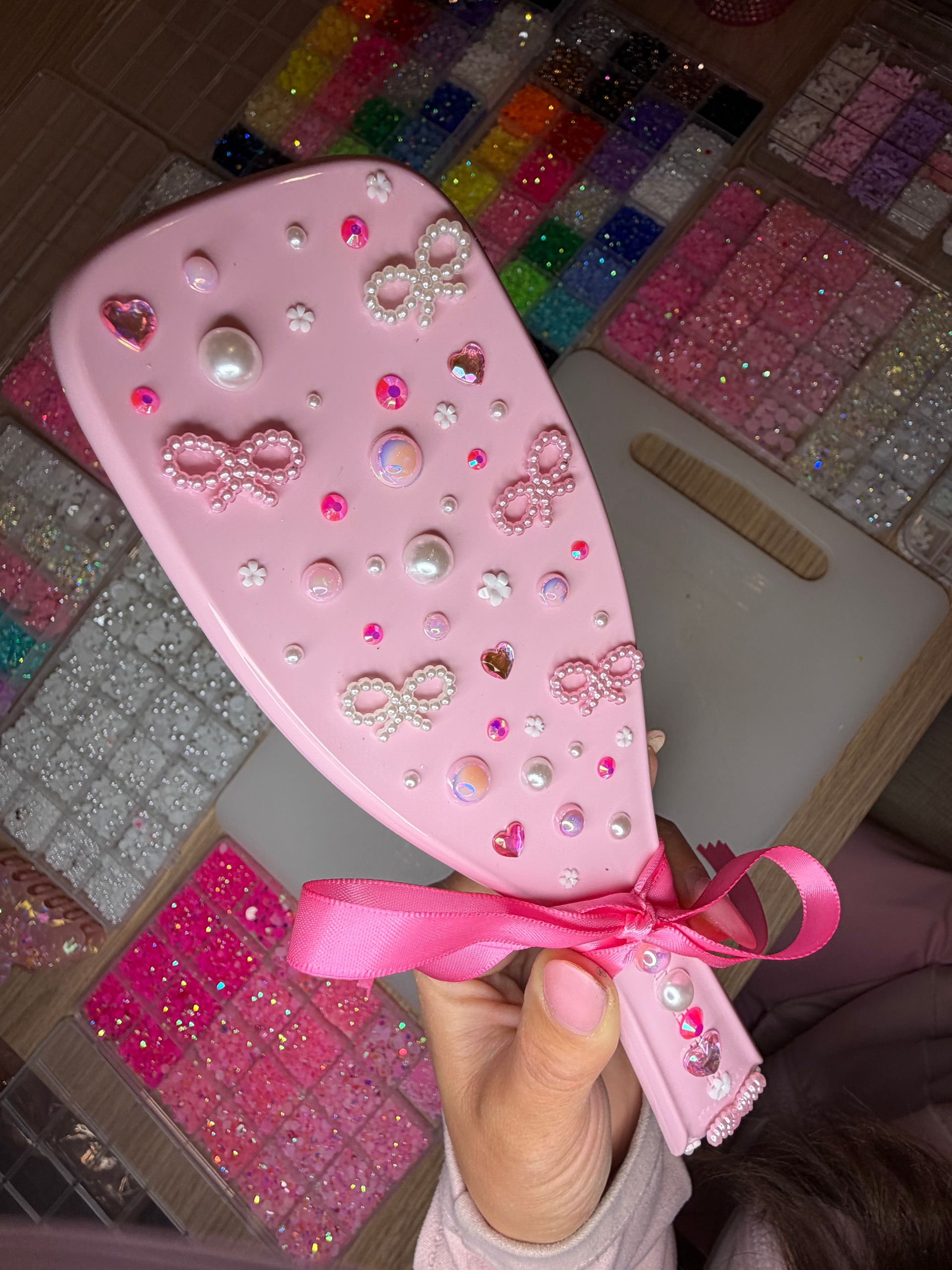 Pink handheld hairbrush decorated with pearl bow embellishments, heart rhinestones, pearls and iridescent gems, held over trays of colorful glitter beads for DIY crafts