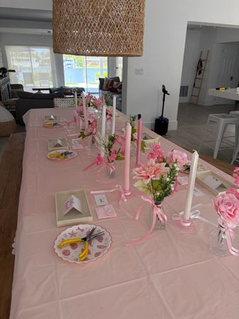 Cheerful pink-themed DIY tablescape in a waterfront open-plan home — long table with pink tablecloth, taper candles, small vases of pink flowers, place cards and craft tools, woven pendant light and canal view.