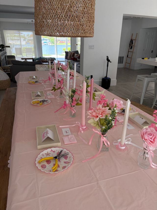 Cheerful pink-themed DIY tablescape in a waterfront open-plan home — long table with pink tablecloth, taper candles, small vases of pink flowers, place cards and craft tools, woven pendant light and canal view.
