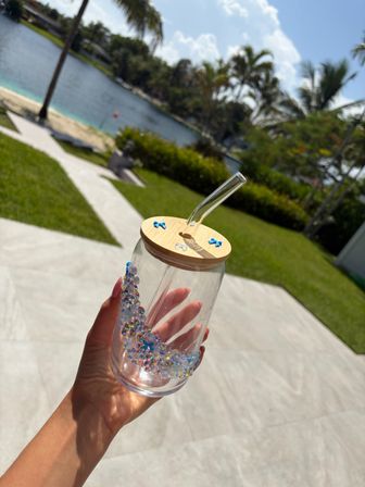 Hand holding a clear glass tumbler with a bamboo lid and angled glass straw, decorated with blue flower accents and iridescent rhinestones, set against a sunny tropical waterfront backyard with palm trees and a paved patio