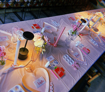 Overhead view of a pink-themed party table set for crafts with heart-shaped plates and place cards, paint palettes, pink candles, glass flutes, floral centerpieces and bar stools by a colorful tiled counter.