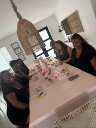 Group of six friends seated at a pink-decorated dining table in a bright modern home, smiling; woven rattan pendant light overhead, pink floral centerpieces, candles and place settings for a celebratory brunch.