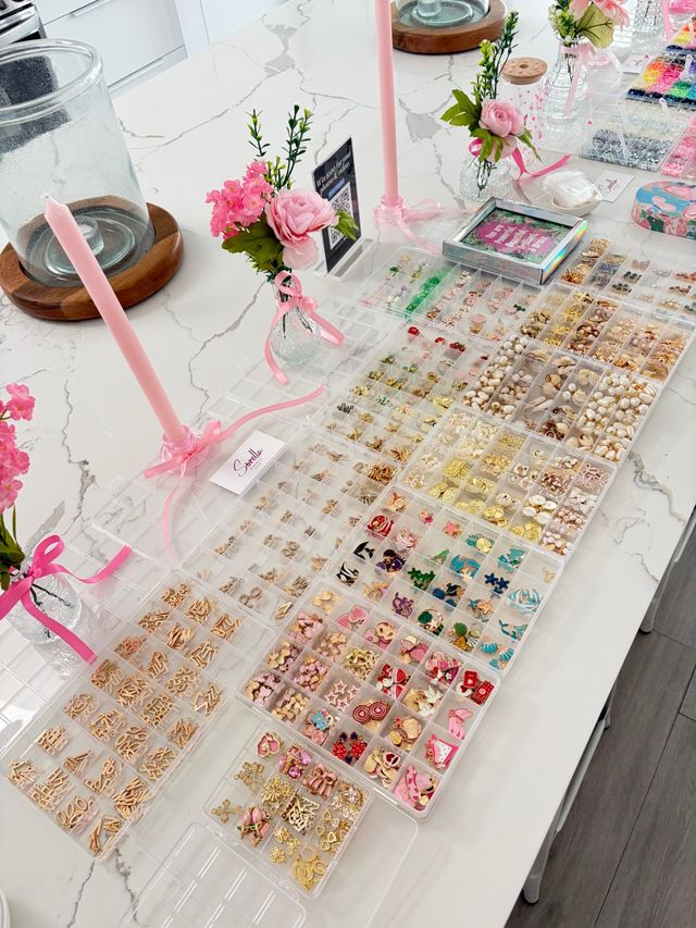 Organized DIY jewelry supplies on a white marble kitchen island: clear compartment trays of gold charms, colorful enamel pieces, pearls and beads, accented by pink taper candles and small floral vases.