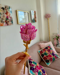 Hand holding a bedazzled pink faux rose with a gold stem in a pastel living room, blush sofa with colorful floral embroidered pillows and framed wall art in the background.