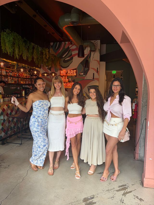Five women in summer outfits smiling and posing under a pink arch at a colorful tropical-themed café bar with hanging plants and a neon sign