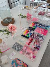 DIY craft station on white marble kitchen island with organized trays of colorful beads, sequins and rhinestones, pink floral accents and candles