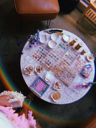 Round terrazzo craft table with jewelry-making supplies: organized trays of tiny gold and colorful charms, shell-shaped dishes of beads and seashells, spools of gold chain, pliers, a purple candle and a small floral vase — beachy beading display.