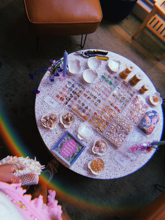 Round terrazzo craft table with jewelry-making supplies: organized trays of tiny gold and colorful charms, shell-shaped dishes of beads and seashells, spools of gold chain, pliers, a purple candle and a small floral vase — beachy beading display.