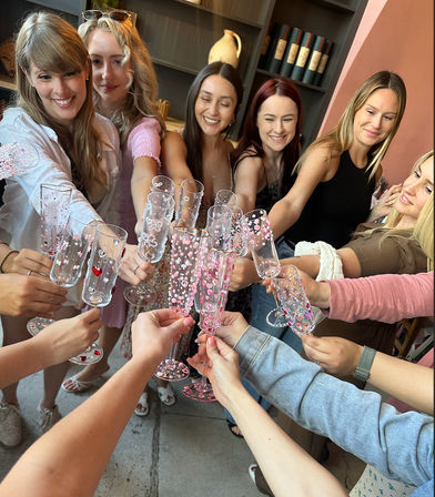 Group of women clinking decorated pink and heart-patterned champagne flutes in a cozy wine-bar or cafe setting — celebratory girls' brunch or night-out toast.