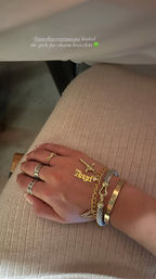 Close-up of a hand on a ribbed beige skirt wearing stacked gold charm bracelets including an Angel nameplate and cross charm, plus multiple rings — stylish layered jewelry at a charm bracelet event.