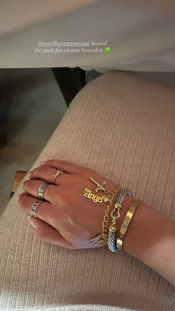 Close-up of a hand on a ribbed beige skirt wearing stacked gold charm bracelets including an Angel nameplate and cross charm, plus multiple rings — stylish layered jewelry at a charm bracelet event.