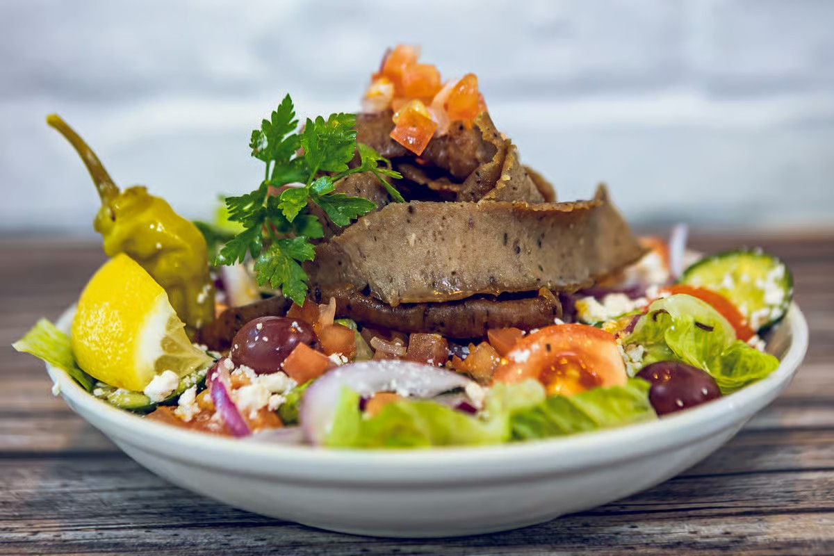 Close-up of a vibrant Mediterranean-style gyro salad: stacked sliced gyro meat over romaine with tomatoes, red onion, Kalamata olives, crumbled feta, lemon wedge, pepperoncini and parsley garnish.