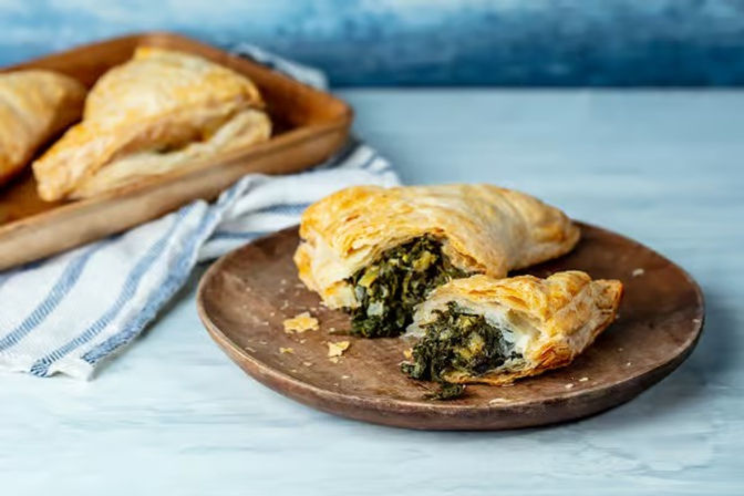 Flaky golden puff pastry turnover split open to reveal vibrant spinach filling on a rustic wooden plate, with a striped kitchen towel and extra turnovers on a tray in the background