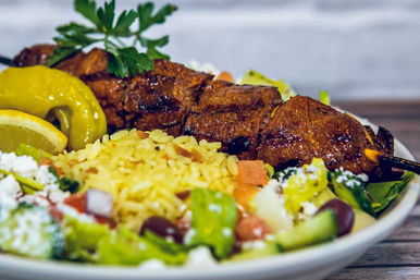 Close-up of Mediterranean-style grilled meat skewer with fragrant yellow rice, lemon wedge, pepperoncini and Greek salad with feta and olives