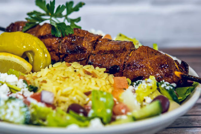Close-up of Mediterranean-style grilled meat skewer with fragrant yellow rice, lemon wedge, pepperoncini and Greek salad with feta and olives