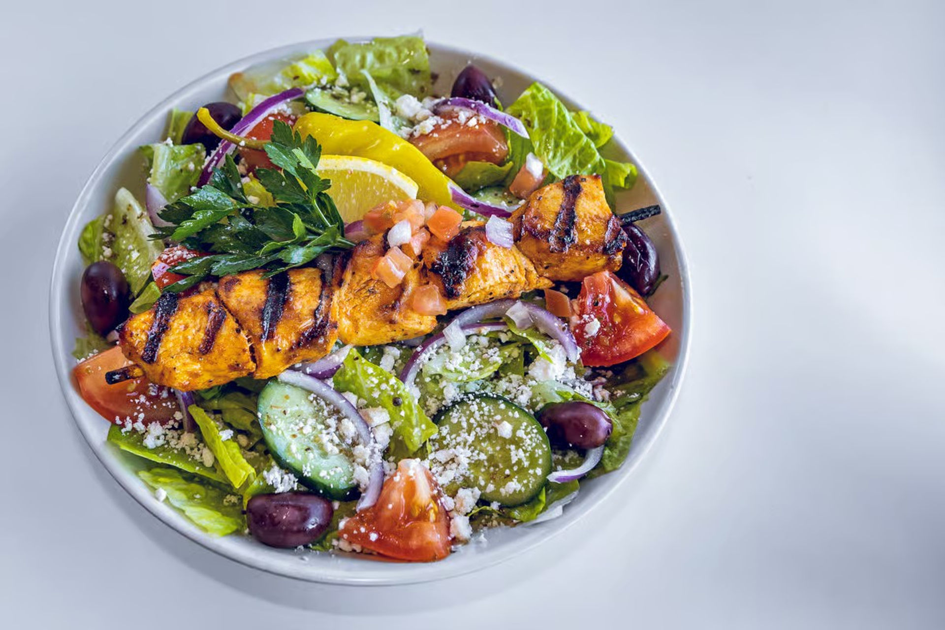 Colorful Greek salad in a white bowl topped with a grilled chicken skewer—romaine, cucumber, tomato, red onion, Kalamata olives, crumbled feta and lemon wedge