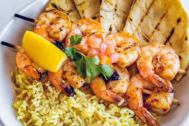 Mediterranean-style seafood bowl with grilled shrimp skewers atop yellow rice, grilled flatbread, lemon wedge, diced tomato and parsley garnish.
