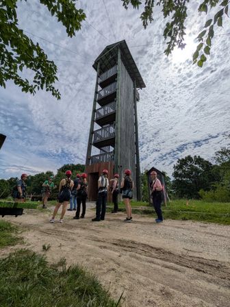 Adventure group in helmets and harnesses gathers at the base of a tall wooden climbing/zipline tower with multi-level platforms and support cables on a grassy, tree‑framed site under a dramatic cloudy sky.