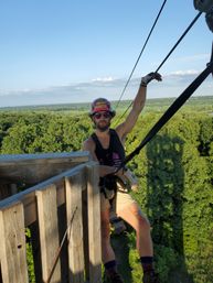 Person in helmet and harness on a wooden tower preparing to zipline over a green forest canopy under a clear blue sky.
