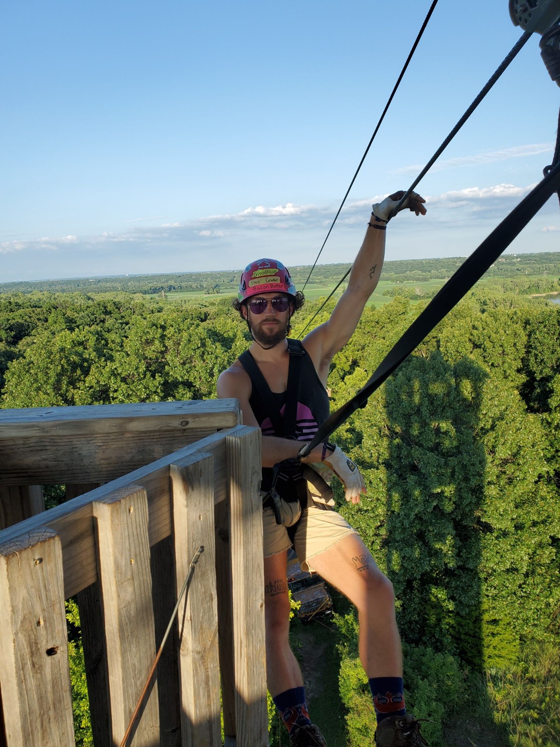 Person in helmet and harness on a wooden tower preparing to zipline over a green forest canopy under a clear blue sky.