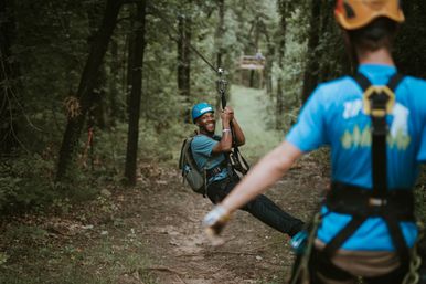 Smiling man in a helmet and backpack gliding on a zipline through a wooded forest trail while a harnessed guide watches — outdoor canopy zipline adventure