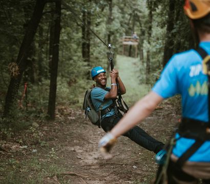 Smiling man in a helmet and backpack gliding on a zipline through a wooded forest trail while a harnessed guide watches — outdoor canopy zipline adventure