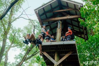 Smiling rider launches from a wooden treetop zipline platform as friends in helmets and harnesses watch amid a green forest canopy - outdoor adventure scene.
