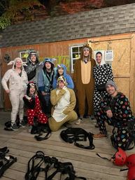 Smiling group of adults wearing animal onesies on a wooden zipline platform at night, surrounded by safety harnesses and red helmets against a rustic shed backdrop.
