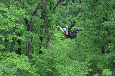 Person wearing a blue helmet and harness ziplining through a dense green forest canopy with arms outstretched