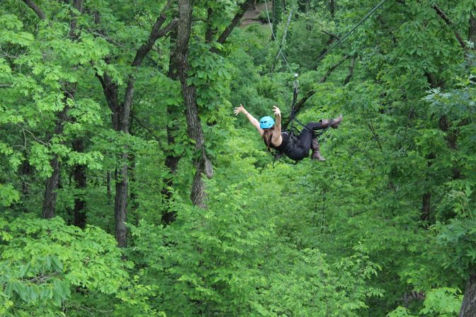 Person wearing a blue helmet and harness ziplining through a dense green forest canopy with arms outstretched