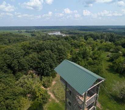 Aerial view from a wooden observation tower with a green metal roof overlooking a lush forest, winding river, grassy clearing with picnic tables, and a wide blue sky dotted with puffy clouds.