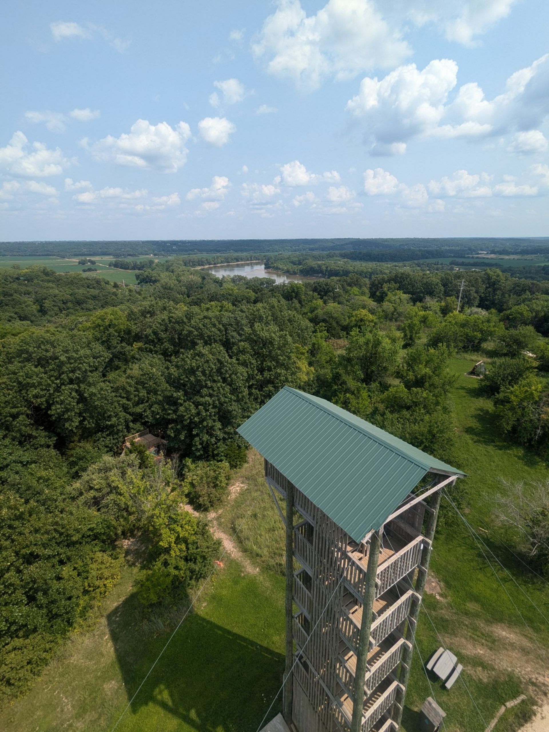 Aerial view from a wooden observation tower with a green metal roof overlooking a lush forest, winding river, grassy clearing with picnic tables, and a wide blue sky dotted with puffy clouds.