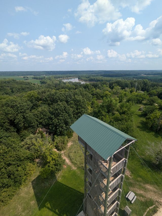 Aerial view from a wooden observation tower with a green metal roof overlooking a lush forest, winding river, grassy clearing with picnic tables, and a wide blue sky dotted with puffy clouds.