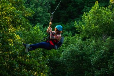 Thrill-seeker in a blue helmet and harness ziplining over a lush green forest canopy with dappled sunlight — outdoor treetop adventure.