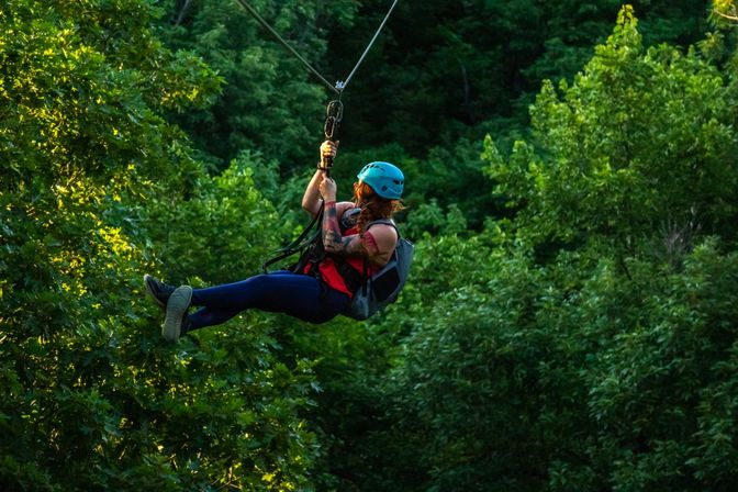 Thrill-seeker in a blue helmet and harness ziplining over a lush green forest canopy with dappled sunlight — outdoor treetop adventure.