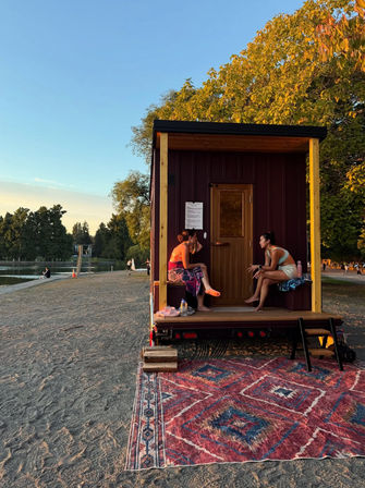 Two women chatting on the porch of a tiny trailer cabin at a lakefront beach park during golden-hour sunset, colorful patterned rug in the foreground and trees and calm water in the background.