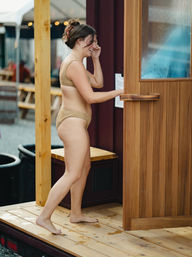 Smiling woman in a tan swimsuit steps barefoot onto a wooden deck and opens a wooden sauna door at an outdoor spa area with picnic tables blurred in the background.