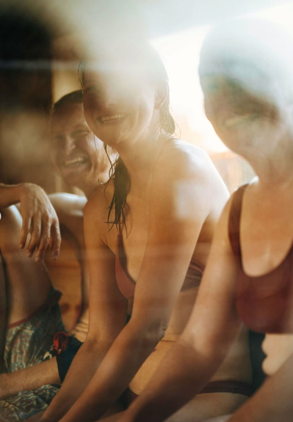 Three smiling people in swimsuits sitting on a wooden bench inside a steamy, sunlit sauna or spa, warm golden light and relaxed atmosphere.