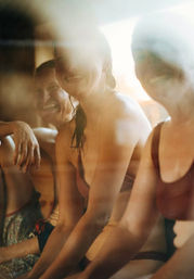 Three smiling people in swimsuits sitting on a wooden bench inside a steamy, sunlit sauna or spa, warm golden light and relaxed atmosphere.