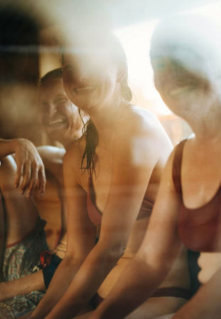 Three smiling people in swimsuits sitting on a wooden bench inside a steamy, sunlit sauna or spa, warm golden light and relaxed atmosphere.