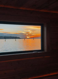 Cozy waterfront sunset viewed through a wooden window, warm orange sky reflecting on calm water with glowing string lights and a distant shoreline silhouette.