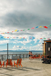 Colorful pennant flags and string lights above a sunny waterfront pier with bright orange wooden chairs, people in swimwear strolling on the boardwalk, calm sea and distant mountains under a partly cloudy sky.