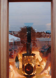 View through a wooden-framed glass door into a warm sauna with a central wood-burning stove, bare feet on wooden benches and an urban waterfront skyline reflected in the glass.