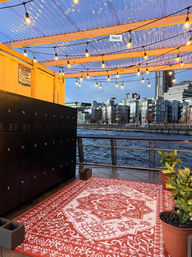 Cozy waterfront patio at twilight with twinkling string lights under a corrugated roof, a red patterned outdoor rug, black lockers, potted plants, and a modern city skyline across the water.