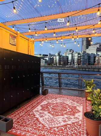 Cozy waterfront patio at twilight with twinkling string lights under a corrugated roof, a red patterned outdoor rug, black lockers, potted plants, and a modern city skyline across the water.